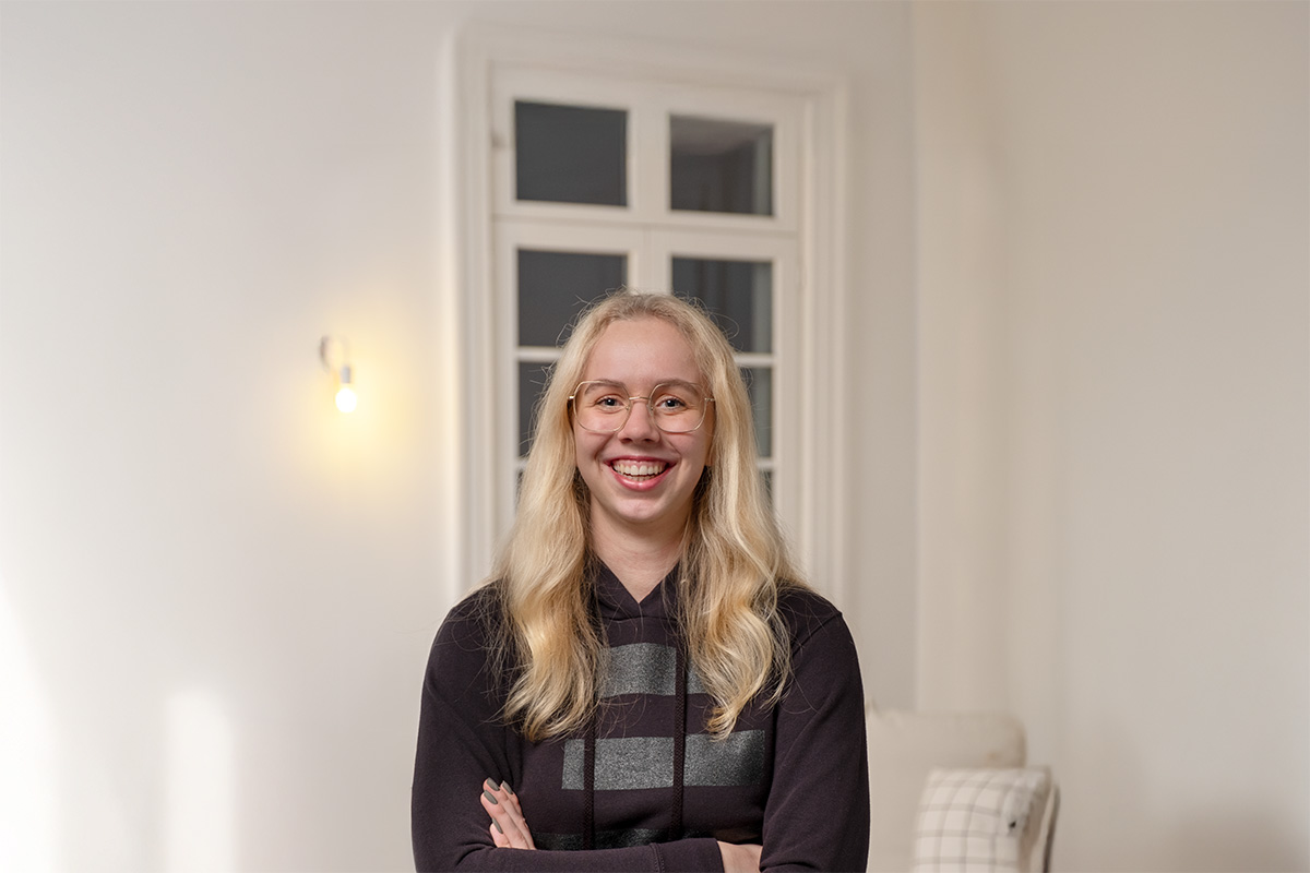 Porträt einer lächelnden Frau mit langen blonden Haaren und Brille, Shayanne Adam, die in einem hellen Raum mit Fenster und Wandlampe steht – Teil des N3W-Messeservice-Teams, das Ausstellern bei der Planung und Umsetzung ihrer Messeauftritte unterstützt.