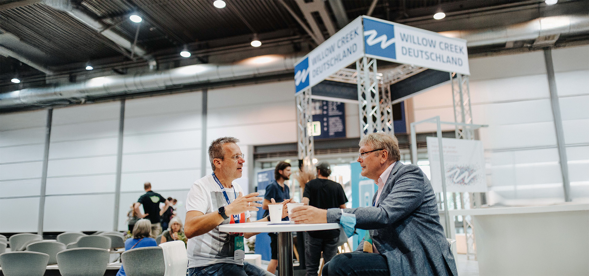 Zwei Männer unterhalten sich an einem Tisch auf einer Messe, im Hintergrund der Stand von Willow Creek Deutschland – ein Beispiel für den professionellen Messeauftritt, den N3W-Messeservice für Aussteller unterstützt.