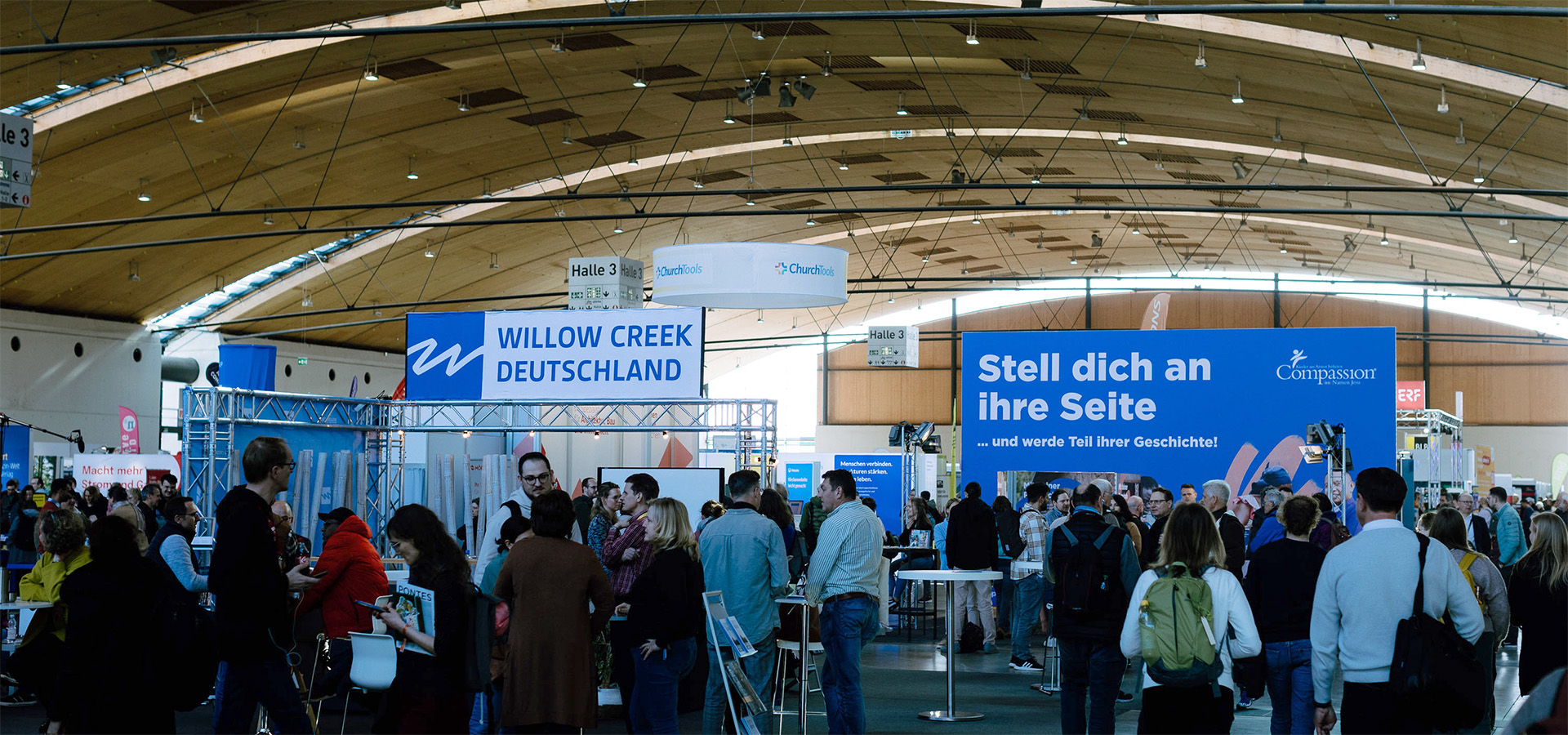Belebte Messehalle mit Ausstellern wie Willow Creek Deutschland und Compassion, wo Besucher netzwerken – N3W-Messeservice unterstützt Aussteller bei Planung, Ausstattung und Umsetzung ihres Messeauftritts für einen professionellen Auftritt.